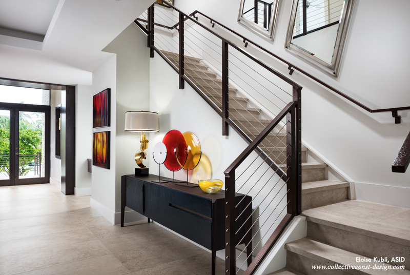 Finished in custom grey satin, LED sconces set off riftcut oak recessed niches. A wood wrap defines the space. The floors and stair case are large-format porcelain concrete slabs. A Moka Oak sideboard with bronze metal details clad in cerulean blue leather anchors the stairway. Three blown-glass spheres from Sweden offset a tall, curled brass lamp and a Murano Glass bowl. Vivid colors are also present in two oils, “Forest Through The Trees”.