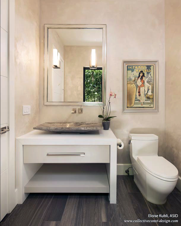 The cabana bath is finished in a light grey lacquer and solid pure white quartz. It is a contrast to the grey carved-stone vessel sink. A silver leaf mirror frame and  mounted lights are a simple and elegant finish that works with the satin nickel wall mount faucet. The walls are burnished with a durable LusterStone® grey finish. The Art Deco framed poster is from the client's personal collection. Cabinetry: KitchenWorks